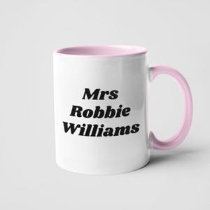 May include: White ceramic mug with a pink rim and handle. The mug has black text that reads "Mrs Robbie Williams".