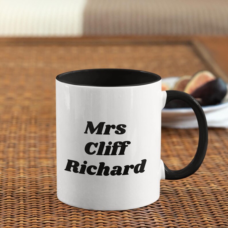 Op de afbeelding: Witte keramische mok met een zwarte rand en handvat. De mok heeft zwarte tekst met de tekst "Mrs Cliff Richard".