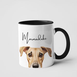 May include: White ceramic mug with a black rim and handle. The mug features a brown and tan dog's face with the name "Marmaduke" written in black cursive.