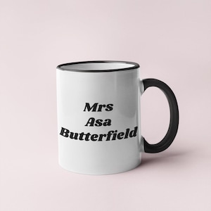 May include: White ceramic mug with a black handle and rim. The mug features the text "Mrs Asa Butterfield" in black, stylized font. The mug is set against a light pink background.