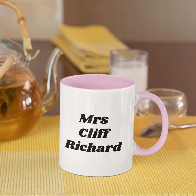 Op de afbeelding: Een witte keramische mok met een roze rand en handvat. De mok heeft zwarte tekst die "Mrs Cliff Richard" luidt.