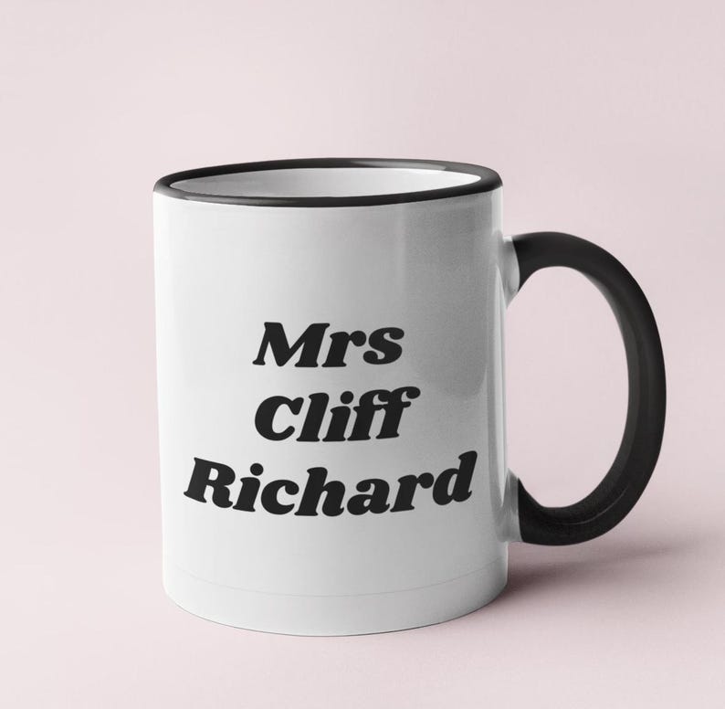Op de afbeelding: Witte keramische mok met een zwarte rand en handvat. De mok heeft zwarte tekst met de tekst "Mrs Cliff Richard".