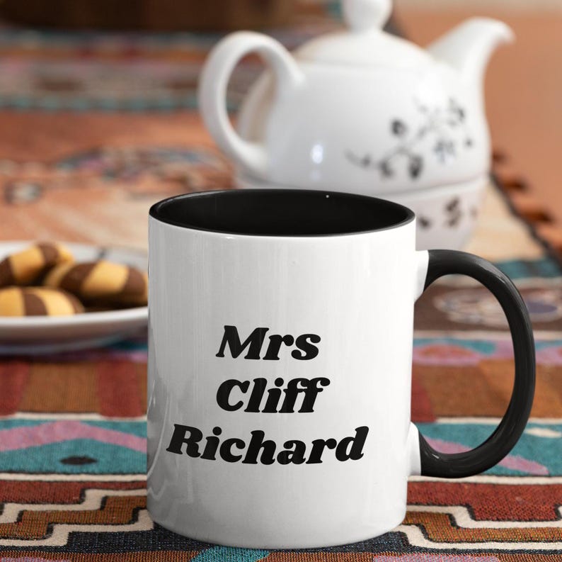 Op de afbeelding: Witte keramische koffiemok met een zwarte rand en handvat. De mok heeft zwarte tekst met de tekst "Mrs Cliff Richard".