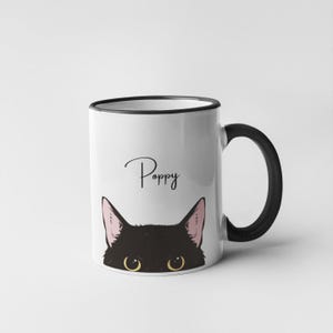 May include: White ceramic mug with a black rim and handle. The mug features a black cat peeking over the top with the name "Poppy" written in black cursive.