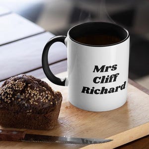 Op de afbeelding: Witte keramische mok met een zwarte rand en handvat. De mok heeft zwarte tekst met de tekst "Mrs Cliff Richard".