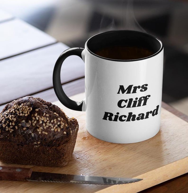 Op de afbeelding: Witte keramische mok met een zwarte rand en handvat. De mok heeft zwarte tekst met de tekst "Mrs Cliff Richard".