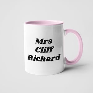 Op de afbeelding: Witte keramische mok met een roze rand en handvat. De mok heeft zwarte tekst die "Mrs Cliff Richard" luidt.