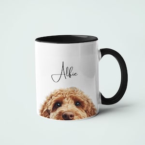 Può includere: Una tazza in ceramica bianca con bordo e manico neri. La tazza presenta un'illustrazione marrone e bianca di un muso di cane che sporge dal bordo. Il nome "Alfie" è scritto in corsivo sulla tazza.