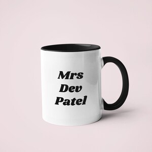Könnte beinhalten: Weiße Keramiktasse mit schwarzem Rand und Henkel. Die Tasse trägt den schwarzen Schriftzug "Mrs Dev Patel".