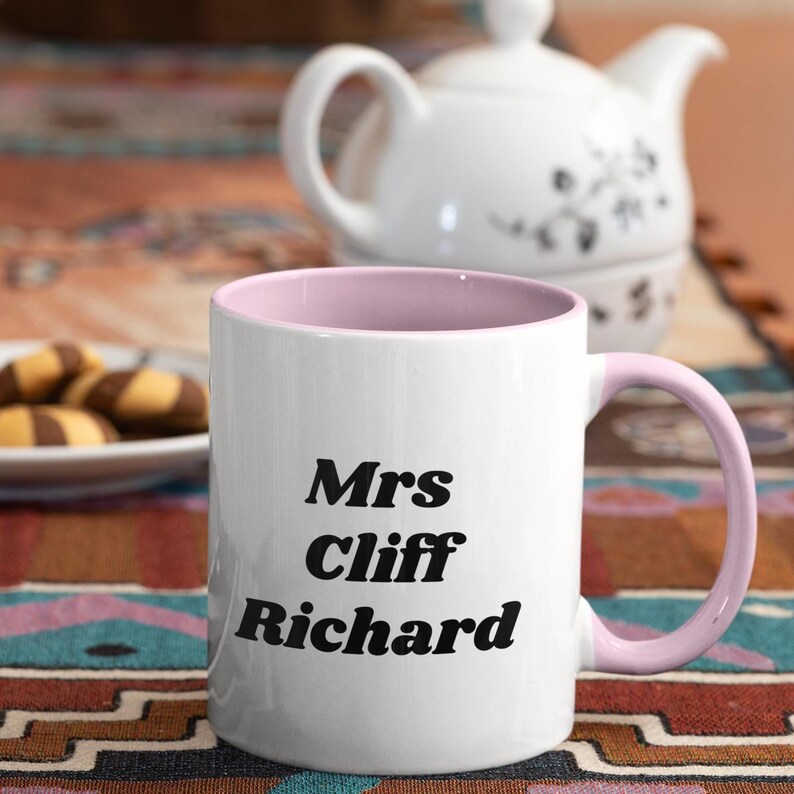 Op de afbeelding: Witte keramische mok met een roze rand en handvat. De mok heeft zwarte tekst met de tekst "Mrs Cliff Richard".