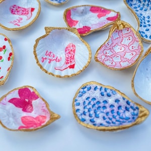 May include: A collection of small, white seashells with gold trim, each decorated with a different colorful pattern. The shells are decorated with designs including cherries, ghosts, boots, and floral patterns. One shell has the text "Let's go girls!" printed on it.