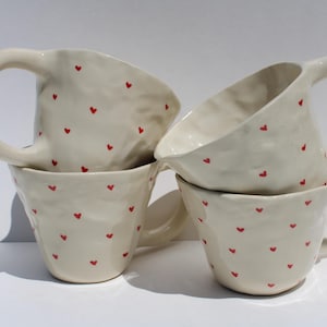 May include: Four off-white ceramic mugs stacked together. Each mug has a large, rounded handle and is decorated with small, red heart shapes. The mugs have a textured surface and a slightly irregular shape.