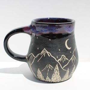 May include: Black ceramic mug with a handle, featuring a mountain and forest design. The mug has a dark blue rim and a crescent moon and stars. The mountain and forest design is in a lighter color, creating a contrast against the black background.