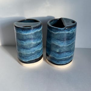 May include: Two ceramic tumblers with a blue, layered design. The cylindrical cups have a black band near the base and a white bottom. One tumbler has a curved opening, the other has a circular opening.