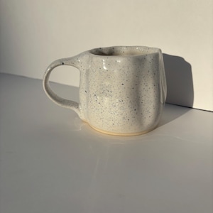 May include: A speckled ceramic mug with a unique, asymmetrical shape. The mug is off-white with dark blue speckles and a rounded handle. The interior and base are a lighter cream color. The mug is designed for beverages.