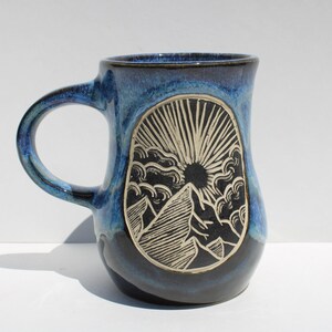 May include: A handmade ceramic mug with a blue glaze. The mug features a mountain and sun design in a black and white oval. The handle is a matching blue. This mug is perfect for coffee, tea, or any hot beverage.