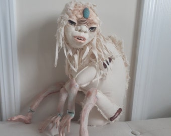 May include: A white fabric doll with a long, white, stringy hair-like fabric. The doll has a light brown face with a turquoise gem on its forehead. The doll has four arms and four hands, all made of fabric.