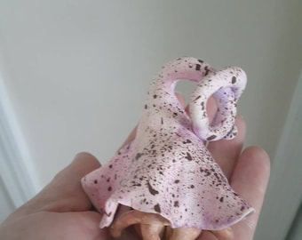 May include: A small, pink and brown ceramic figurine of a fairy with green wings. The fairy is wearing a pink dress with brown spots and has her arms crossed.