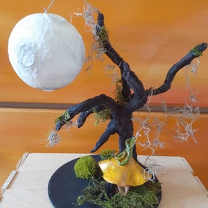 May include: A whimsical miniature tree with a paper mache moon hanging above a yellow mushroom figurine. The tree is made of driftwood and has moss growing on it. The base is a black circular plate with moss and a small green moss ball.