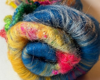 May include: A tightly wound ball of colorful fiber. The yarn features a blend of vibrant colors, including blue, yellow, pink, and green, with strands of silver metallic thread. The texture appears soft and fluffy, suitable for various craft projects.
