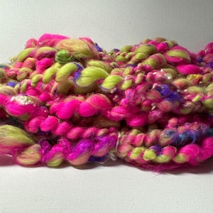Può includere: Un mix di filati colorati con sfumature di rosa, verde e viola. Il filato è filato a mano e ha un aspetto soffice e testurizzato.