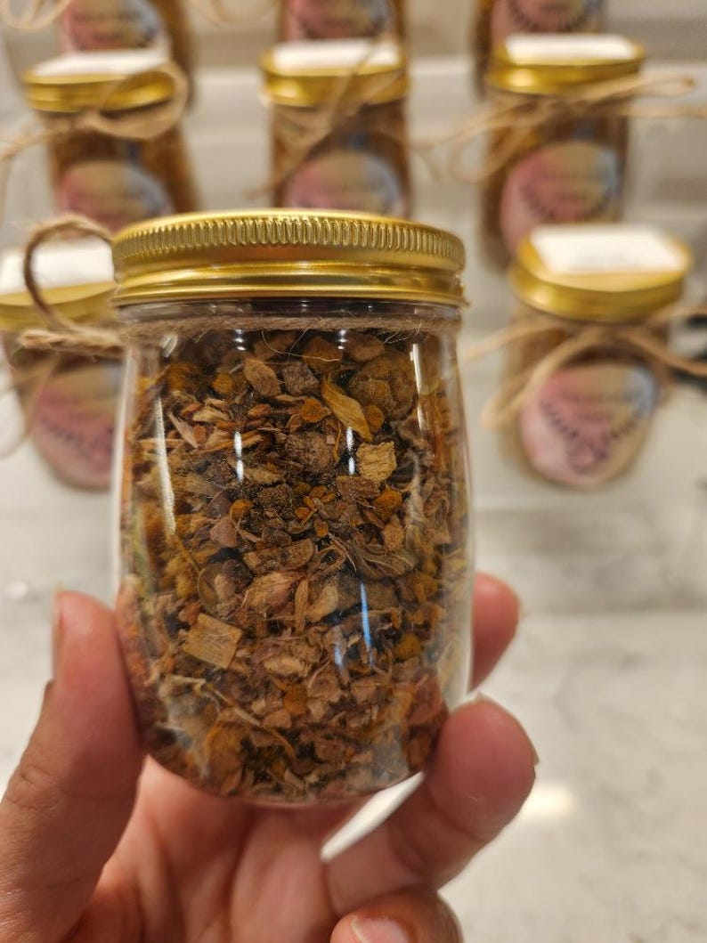 Puede incluir: Un frasco de vidrio con tapa dorada lleno de hierbas y flores secas. El frasco est&aacute; sobre una mesa con otros frascos.
