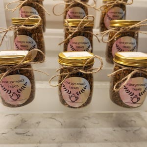 Puede incluir: Nueve frascos de vidrio con tapas doradas llenos de una mezcla marr&oacute;n. Cada frasco tiene una etiqueta blanca con el texto "Love Joy Peace Treats" y una ilustraci&oacute;n rosa y blanca de una tetera con un dise&ntilde;o floral.
