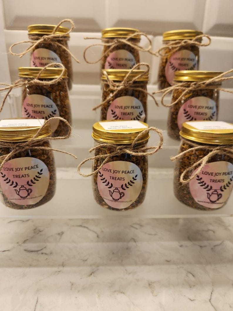 Puede incluir: Nueve frascos de vidrio con tapas doradas llenos de una mezcla marr&oacute;n. Cada frasco tiene una etiqueta blanca con el texto "Love Joy Peace Treats" y una ilustraci&oacute;n rosa y blanca de una tetera con un dise&ntilde;o floral.