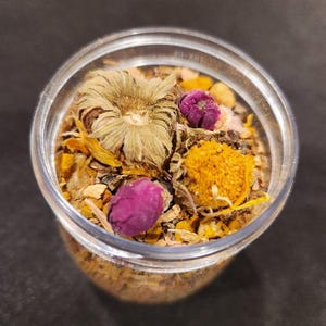 Puede incluir: Un tarro de vidrio transparente lleno de una mezcla de hierbas y flores secas. La mezcla incluye flores amarillas secas, flores moradas secas y flores marrones secas.