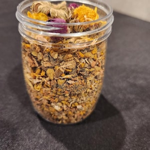 Puede incluir: Un tarro de vidrio transparente lleno de hierbas y flores secas, incluyendo flores amarillas, naranjas y moradas. El tarro est&aacute; sobre una superficie negra.