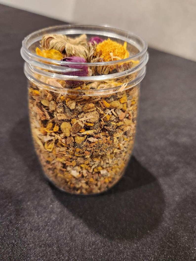 Puede incluir: Un tarro de vidrio transparente lleno de hierbas y flores secas, incluyendo flores amarillas, naranjas y moradas. El tarro est&aacute; sobre una superficie negra.