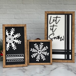 May include: Three rectangular and square wooden framed signs with a natural wood finish. Two signs feature white snowflake cutouts with Mickey Mouse icon details on a black background. One sign features the black text Let it Snow on white.