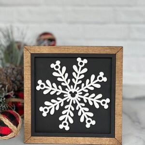 May include: A square wooden sign with a natural wood frame and a black background. Centered on the sign is a white snowflake cutout. The snowflake features six symmetrical branches, each ending in a three-circle icon, with a matching icon at the center.
