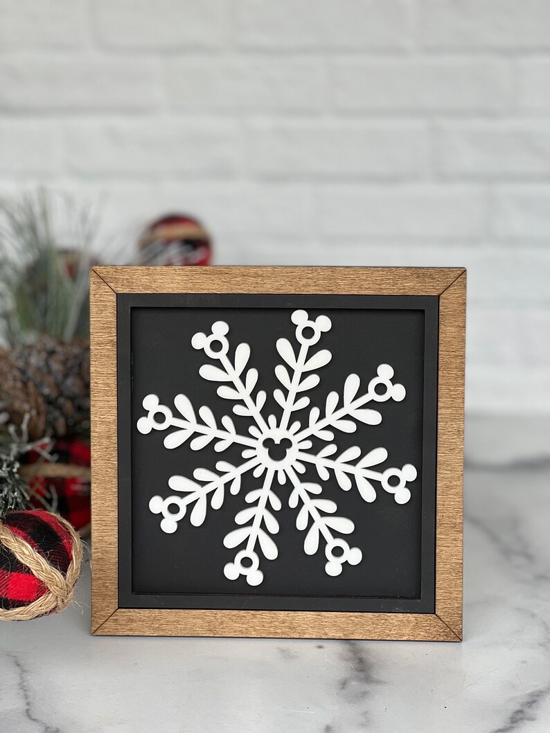 May include: A square wooden sign with a natural wood frame and a black background. Centered on the sign is a white snowflake cutout. The snowflake features six symmetrical branches, each ending in a three-circle icon, with a matching icon at the center.