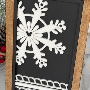 May include: A rectangular wall sign featuring a light brown wooden frame and a black background. A white, layered snowflake cutout with small circular mouse-ear shapes at the tips is centered, with two horizontal white rope-like borders at the bottom.
