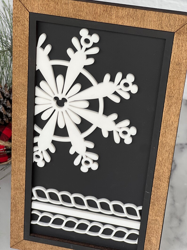 May include: A rectangular wall sign featuring a light brown wooden frame and a black background. A white, layered snowflake cutout with small circular mouse-ear shapes at the tips is centered, with two horizontal white rope-like borders at the bottom.