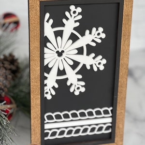 May include: A rectangular wooden sign with a dark brown frame and a black background. The sign features a white cutout of a snowflake with small mouse-ear shapes at the tips and center, and a white rope-like border design at the bottom.