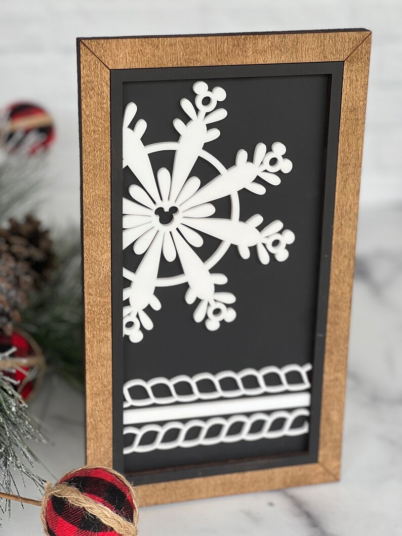 May include: A rectangular wooden sign with a dark brown frame and a black background. The sign features a white cutout of a snowflake with small mouse-ear shapes at the tips and center, and a white rope-like border design at the bottom.