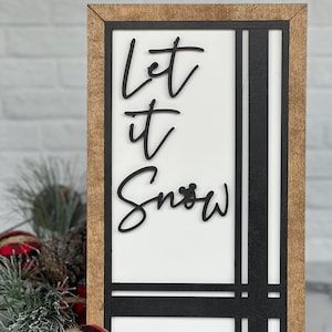 May include: A rectangular wooden sign with a light brown frame and a white center panel. The sign features black, raised lettering that reads Let it Snow, with a small mouse ear icon integrated into the letter o. Black vertical and horizontal lines accent the right and bottom edges of the white panel.