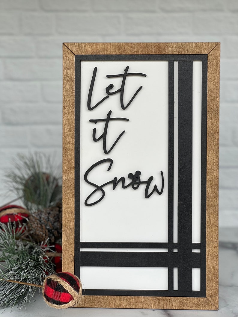 May include: A rectangular wooden sign with a light brown frame and a white center panel. The sign features black, raised lettering that reads Let it Snow, with a small mouse ear icon integrated into the letter o. Black vertical and horizontal lines accent the right and bottom edges of the white panel.