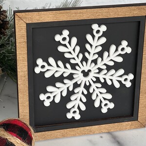 May include: A square wooden sign featuring a white snowflake cutout centered on a black background. The snowflake has six arms, each ending in a small circular icon resembling a mouse head, with a matching icon at the center of the design.