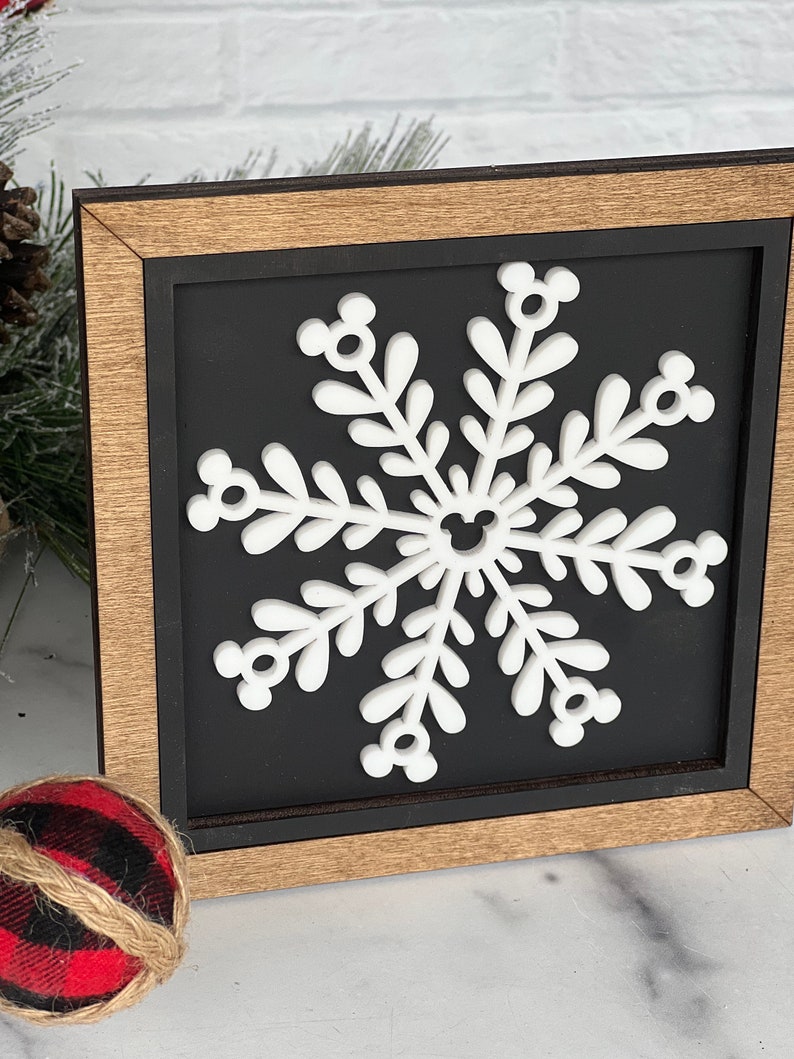 May include: A square wooden sign featuring a white snowflake cutout centered on a black background. The snowflake has six arms, each ending in a small circular icon resembling a mouse head, with a matching icon at the center of the design.