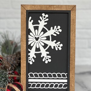 May include: A rectangular wooden sign with a dark brown frame and a black background. The sign features a white cutout of a partial snowflake with Mickey Mouse icon details and two horizontal white rope-patterned borders at the bottom.
