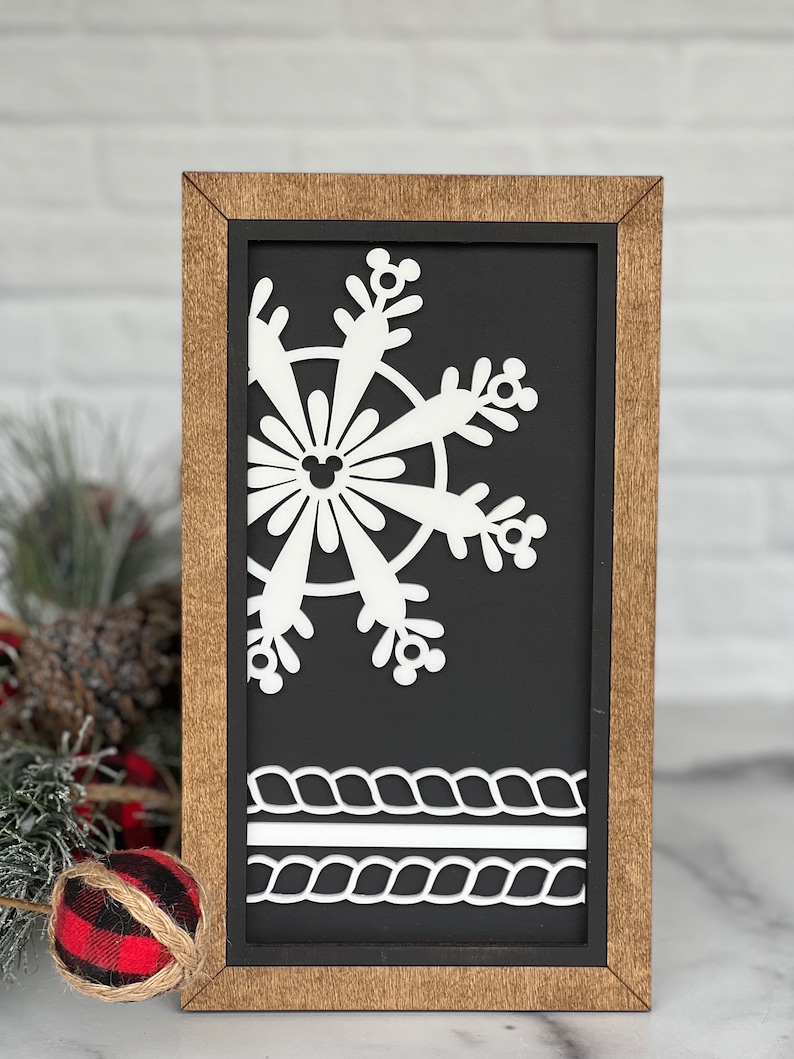 May include: A rectangular wooden sign with a dark brown frame and a black background. The sign features a white cutout of a partial snowflake with Mickey Mouse icon details and two horizontal white rope-patterned borders at the bottom.