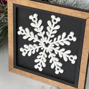 May include: A square wooden sign with a natural wood frame and a black background. Centered on the sign is a white, three-dimensional snowflake cutout. The snowflake features six arms, each ending in a small circular icon resembling a mouse head.