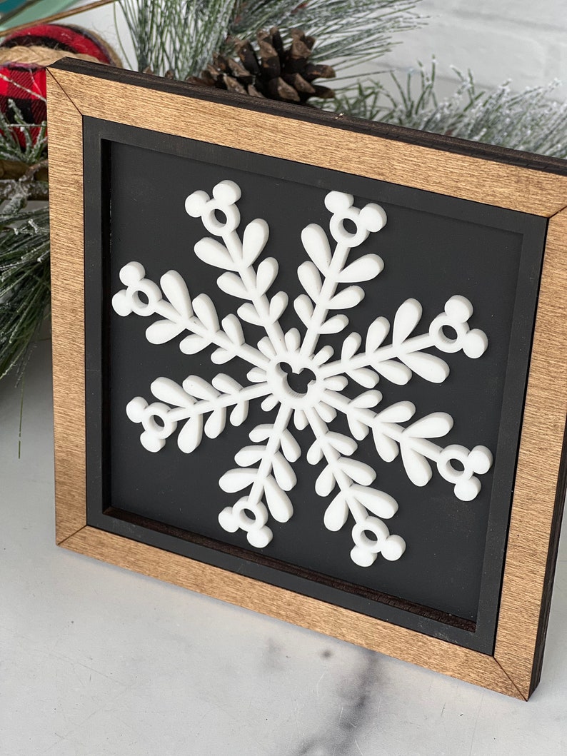 May include: A square wooden sign with a natural wood frame and a black background. Centered on the sign is a white, three-dimensional snowflake cutout. The snowflake features six arms, each ending in a small circular icon resembling a mouse head.