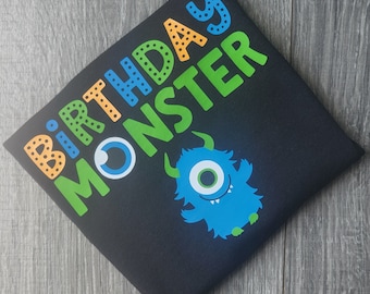 May include: Black t-shirt with the text "Birthday Monster" in green, orange, and blue. A blue monster with a single eye is printed on the shirt.