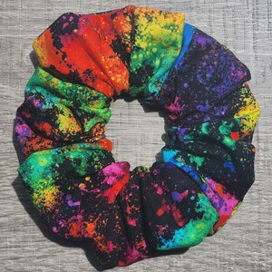 May include: A colorful hair scrunchie with a black background and a splatter paint design in shades of red, orange, yellow, green, blue, and purple.