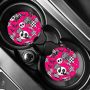 May include: Two car coasters with a pink background and a black and white pattern of skulls, roses, bats, stars, and checkered hearts. The coasters are in a car's cup holders.