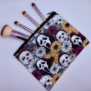 Può includere: Un beauty case con cerniera nera, con un motivo floreale con personaggi di film horror. Sono visibili diversi pennelli per il trucco con manici rosa e setole beige. Il design della borsa include immagini di Michael Myers, Ghostface e Jason Voorhees.